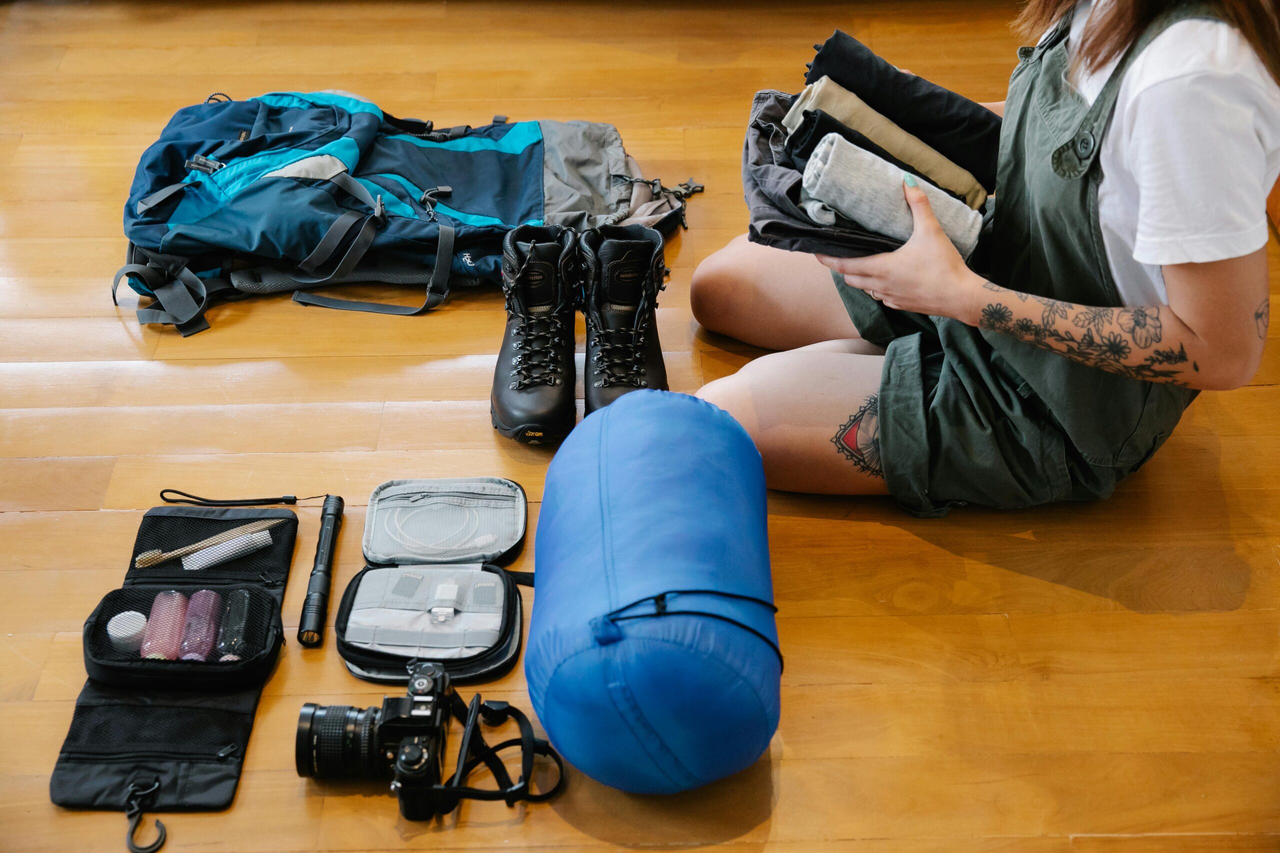 Woman organizing camping gear and clothes on wooden floor indoors, preparing for an adventure.